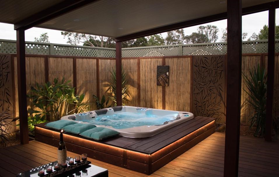 A spa pool set into a wooden deck, surrounded by cushioned seating. Beautiful spa landscaping.